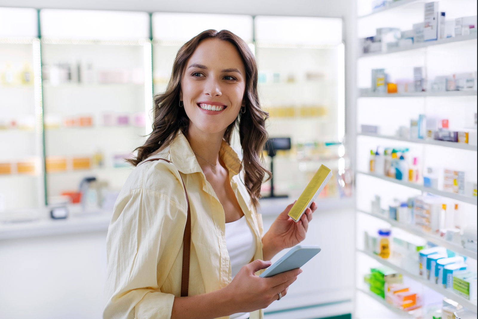 Frau mit Handy steht in der Apotheke und hält ein Medikament in der Hand